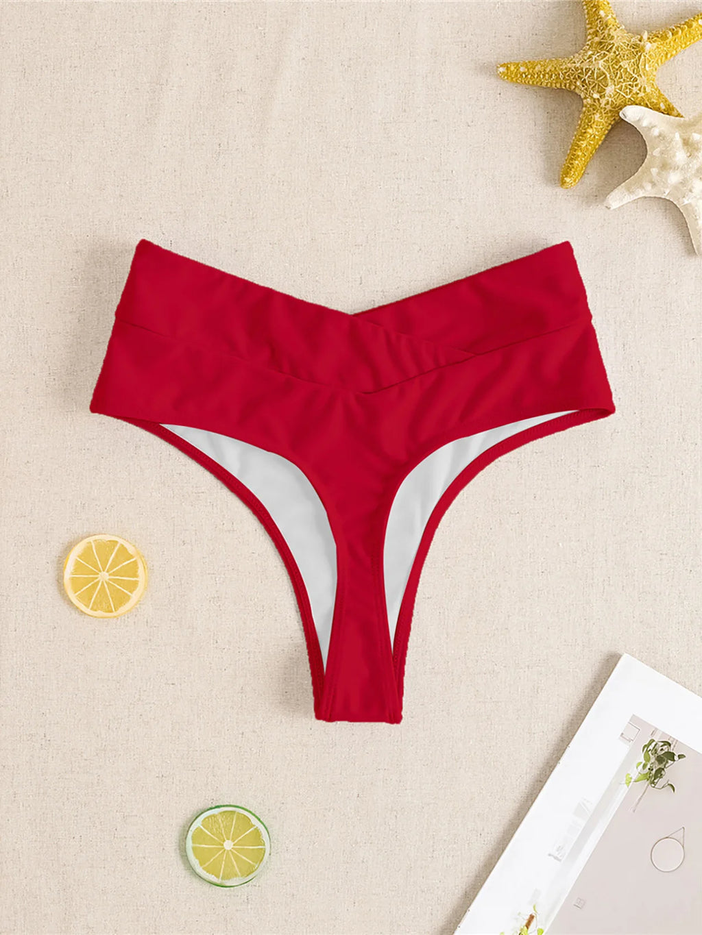 Red high waist crossed waistband Brazilian bikini bottom flat on beige linen with starfish props