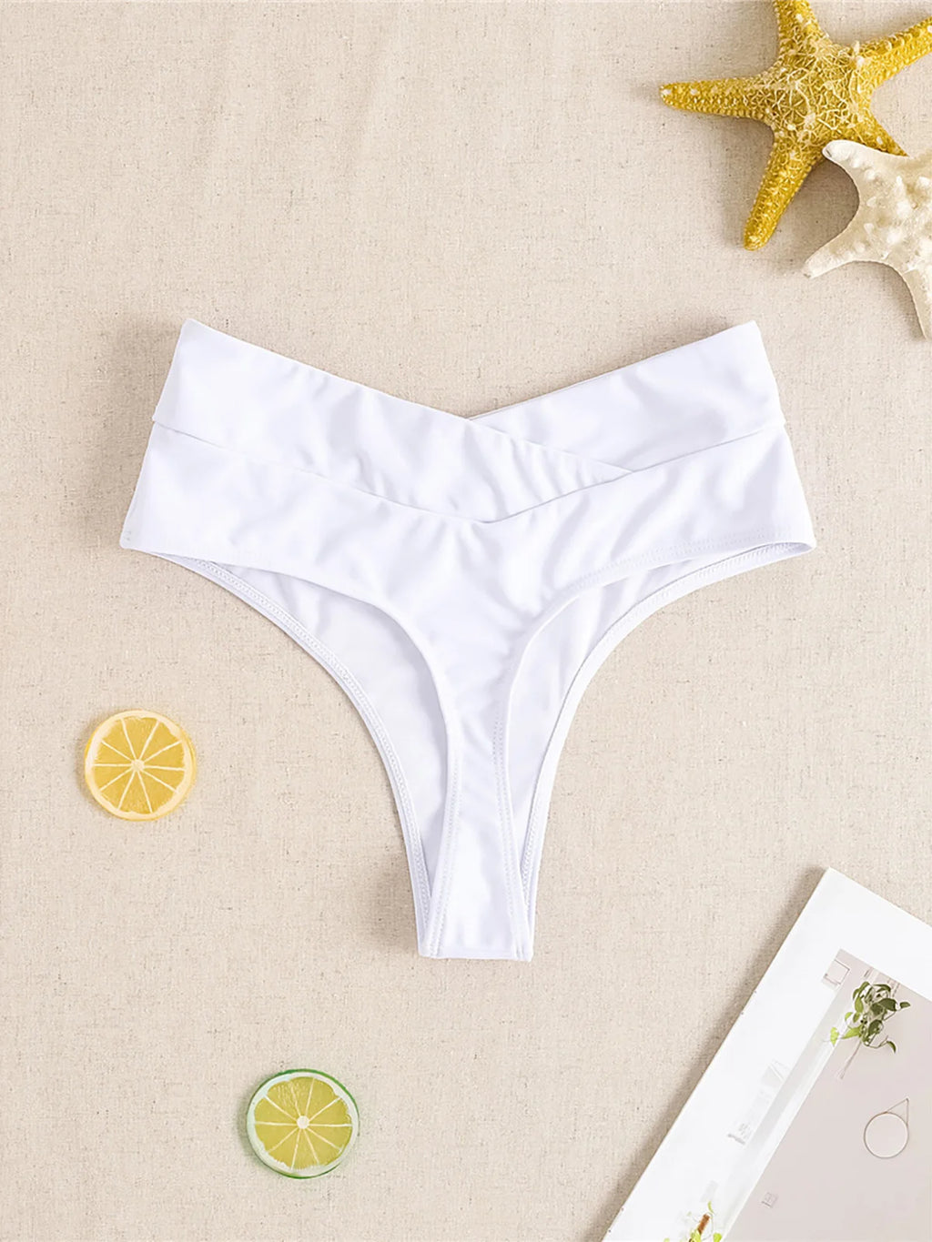 White high waist crossed waistband Brazilian bikini bottom flat on beige linen with starfish props