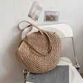 Woven straw round tote with tan leather strap, laid on a white tabletop